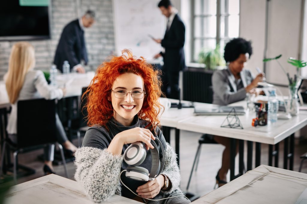 Young happy redhead businesswoman holding headphones and looking at camera while sitting at her desk in the office. Her colleagues are working in the background.