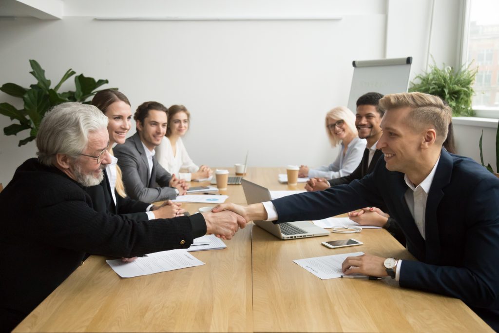 Meeting Room Planning: Young and old male partners shaking hands after signing contracts at group multiracial meeting, senior investor buying startup promising support handshaking entrepreneur, partnership deal concept